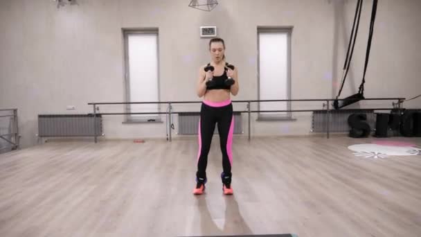 young slim woman in sportswear jumps and half squats with dumbbells against white windows in gym - Záběry, video