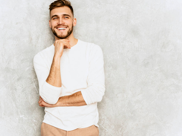 Πορτραίτο του όμορφος χαμογελώντας hipster lumbersexual επιχειρηματίας μοντέλο φοράει ρούχα casual καλοκαιρινό λευκό. Μόδα κομψό άνδρα θέτοντας γκρίζο τοίχο. Αγγίζοντας το πηγούνι και σκέψης - Φωτογραφία, εικόνα
