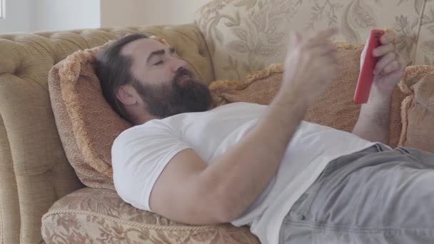 Handsome bearded man lying on the sofa checking cell phone. Guy is angry, he throws away smartphone. Receiving bad news. Emotional man rests at home - Séquence, vidéo