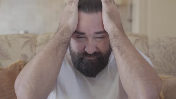Portrait of bearded massaging his head with big hands with expression of suffering on face close up. Adult man has strong headache pain sitting on couch at home - Séquence, vidéo