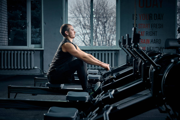 Hombre joven fuerte en una camiseta y pantalones cortos haciendo ejercicios en los simuladores en el gimnasio mirando a un lado
 - Foto, Imagen