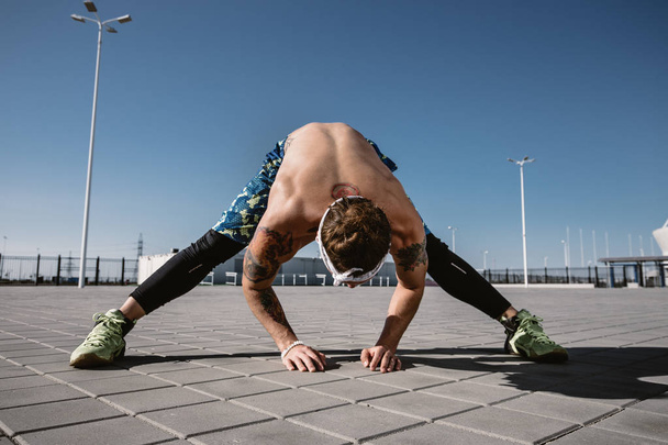 Handsome shirtless sportsman looking away doing stretching exercise on workout ground outdoors at bright sunny day. Sport lifestyle - Foto, afbeelding