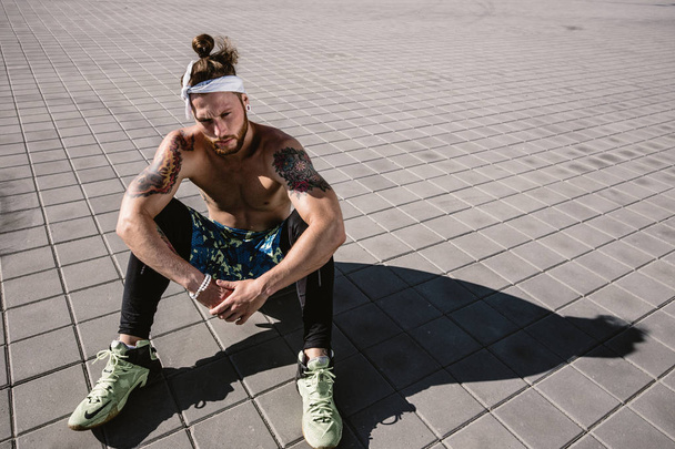 Young  athletic man with a naked torso with headband and tattoos dressed in the black leggings and blue shorts sits on paving slabs on a warm sunny day - Φωτογραφία, εικόνα