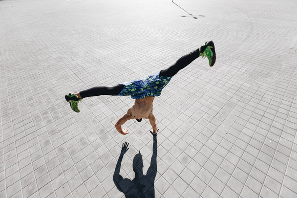 Athletic guy with a naked torso with tattoos dressed in the  black leggings and blue shorts doing the trick standing on one hand on the paving slabs in the street - 写真・画像