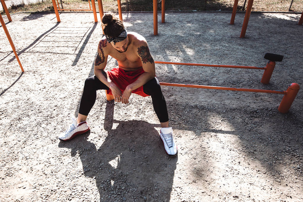 Young athletic man in headband with naked torso dressed in black leggings and red shorts sits near the sport equipment on the sports ground with sand on a sunny day - Фото, зображення