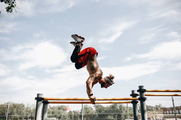 Young athletic man in headband with naked torso dressed in black leggings and red shorts doing a handstand on uneven bars on the sports ground outside on a sunny day - Fotó, kép