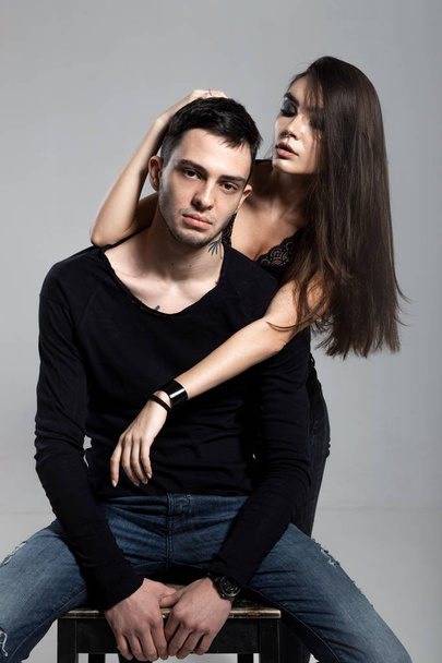 Beautiful brunette girl hugging from behind a guy dressed in a black long sleeve t-shirt and jeans sitting on a wooden chair. Young and cocky lovers - Zdjęcie, obraz
