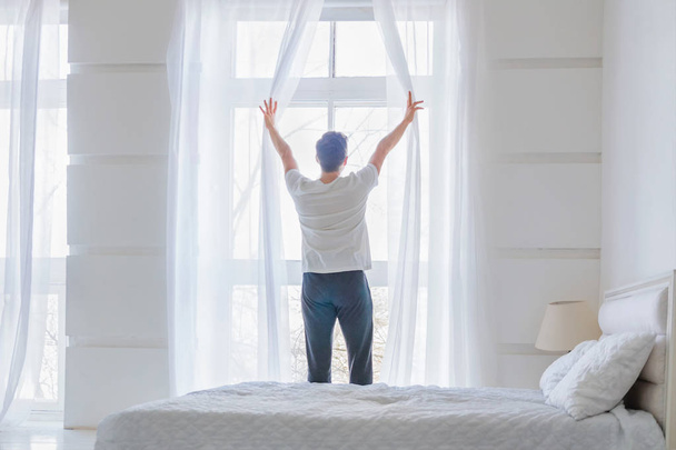 Młody człowiek w pobliżu okna w domu. Otworzenie zasłony w streszczenie sypialnia biały człowiek - Zdjęcie, obraz