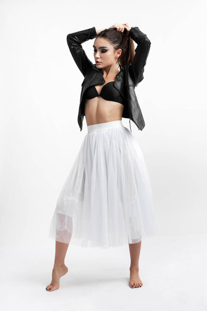 Beautiful brunette girl in a white tulle skirt in a black bra and black leather jacket stands on the white background in the studio - Φωτογραφία, εικόνα
