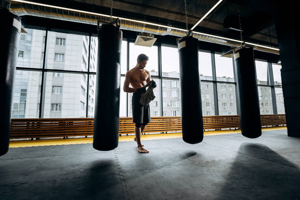 Guy with a naked torso dressed in the black shorts stands next to the punching bags against the background of panoramic windows in the gym - 写真・画像