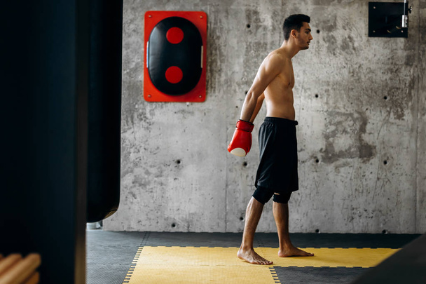 Βάναυση μελαχρινός άντρας με ένα γυμνό κορμό και τα γάντια του μποξ κόκκινο αντιπροσωπεύει κατά ένα συγκεκριμένο τοίχο δίπλα στον εξοπλισμό για την πυγμαχία - Φωτογραφία, εικόνα