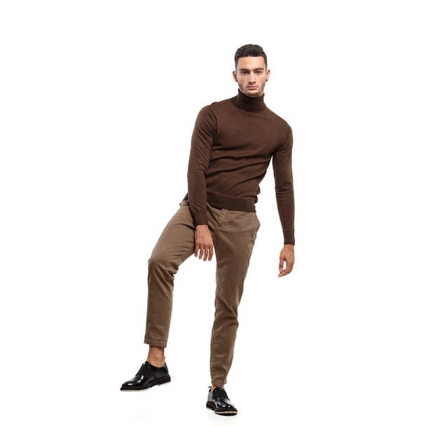 Young stylish man dressed in brown turtleneck and pants stands on the white background in the studio - Foto, Bild