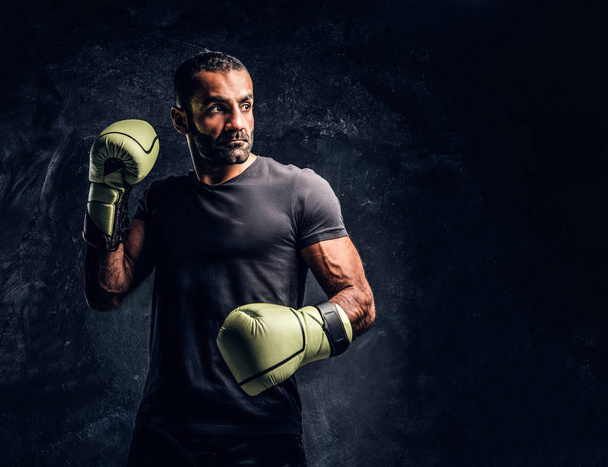 Portrait of a brutal professional fighter in a black shirt and gloves. Studio photo against a dark textured wall - Fotoğraf, Görsel