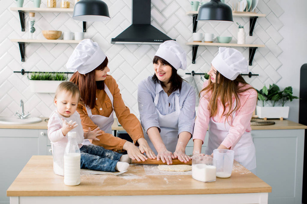 Smiling middle aged woman in kitchen apron rolling out dough and two daughters helping her. Litle baby girl sitting on the table and having fun. Happy women in white aprons baking together - Φωτογραφία, εικόνα