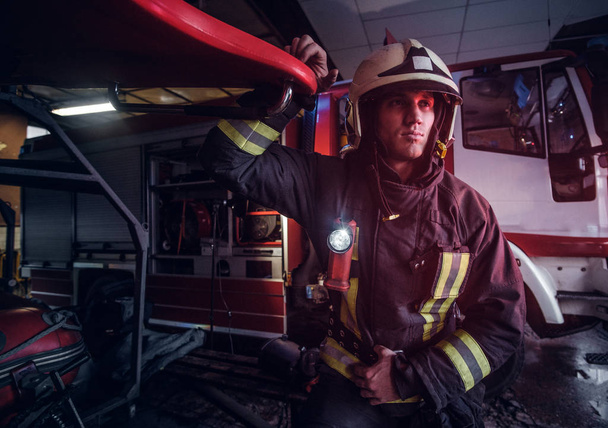 Portrait d'un beau pompier portant un uniforme de protection avec lampe de poche inclus debout dans un garage de la caserne de pompiers
 - Photo, image