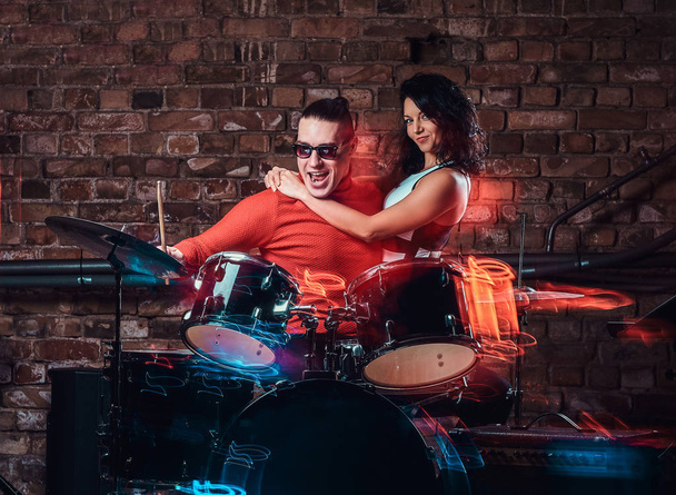 Девушка обнимает своего кумира, который играет на барабане в ночном клубе у кирпичной стены
 - Фото, изображение