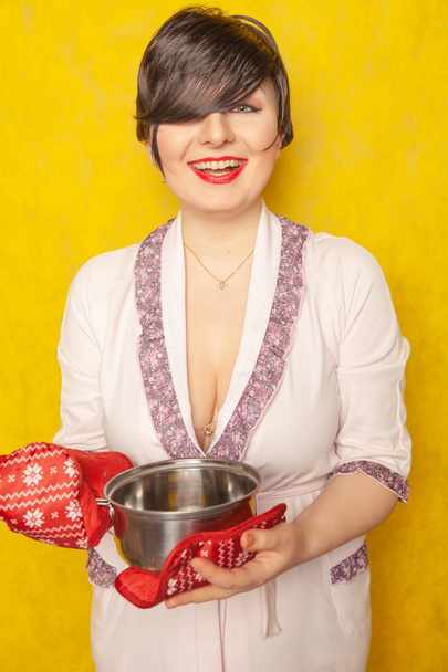 Kavkazská hospodyně připraví jídlo. brunetka stojí v županu s pánev a hrnec držáky na žlutém pozadí solidní Studio. - Fotografie, Obrázek