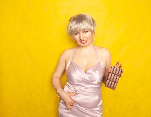 肉付きの良い女性彼女の手で不幸な顔とチョコレートバーの略で、黄色固体のスタジオの背景に彼女のぽっちゃり腹の脂肪に触れる - 写真・画像