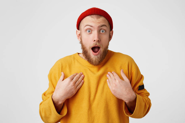 Amazed male keeps mouth opened, hands on breast, being shocked, stares at camera, isolated over white background. Bearded man in red hat man expresses great surprisement - Photo, image