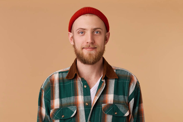 Attractive young man in studio looking at camera. Portrait of a normal guy with broad in red hat and checkered shirt in front of beige background - Fotoğraf, Görsel