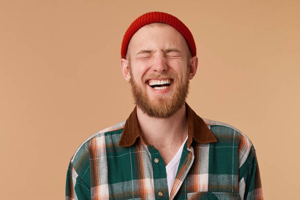 Laughing man in checkered shirt and red hat over beige wall. Toothy smile and beard. Male laughing out loud at funny meme he found on internet. Positive human facial expressions and emotions - Foto, Imagen