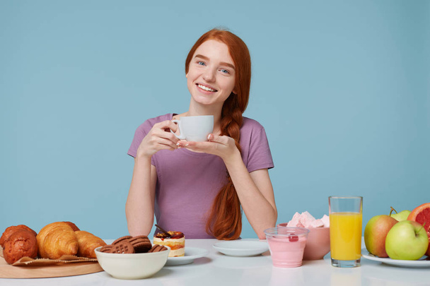 Red-haired girl sitting at a table with slightly bowed head, smiles holds white cup with delicious drink in hands, has lunch looking camera. On the table baking products and fresh food lay - Foto, Bild