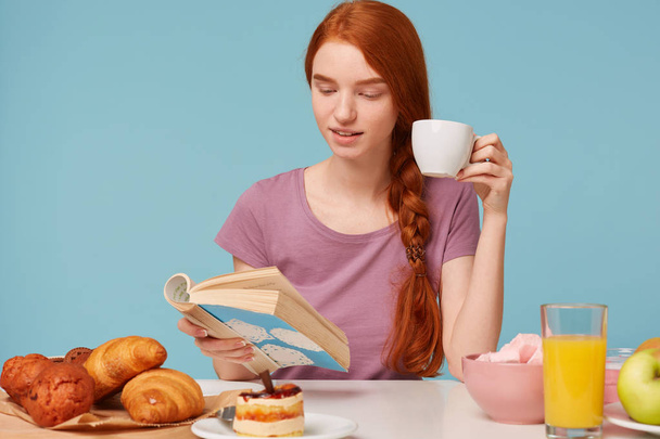 Крупным планом очаровательная рыжая девушка с плетеными волосами, сидящая за столом, пьет из белой чашки вкусный чай, завтракает чтением книги. На столе хлебобулочные изделия и свежие продукты лежали, над синей стеной
 - Фото, изображение