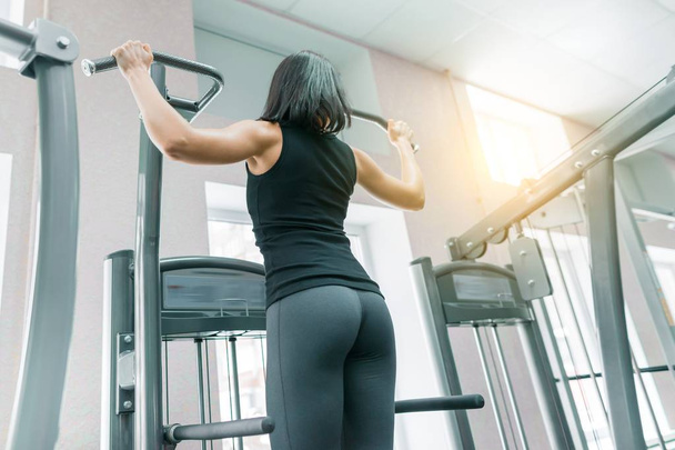 Young woman doing exercises for the back on fitness machine in gym, view from back. Fitness, sport, training, people concept. - 写真・画像