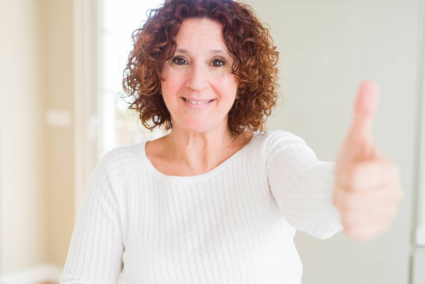 Bella donna anziana indossa maglione bianco a casa facendo pollici felici gesto con la mano. Approvazione espressione guardando la fotocamera che mostra il successo
. - Foto, immagini