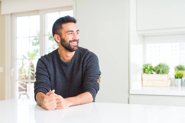 Hombre hispano guapo usando suéter casual en casa mirando hacia otro lado con sonrisa en la cara, expresión natural. Riendo confiado
. - Foto, imagen