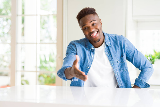 Hombre afroamericano guapo en casa sonriendo amistoso ofreciendo apretón de manos como saludo y bienvenida. Negocios exitosos
. - Foto, Imagen