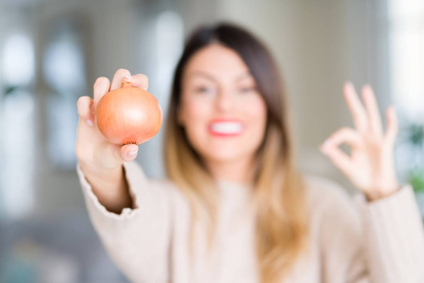 Young beautiful woman holding fresh onion at home doing ok sign with fingers, excellent symbol - Photo, Image