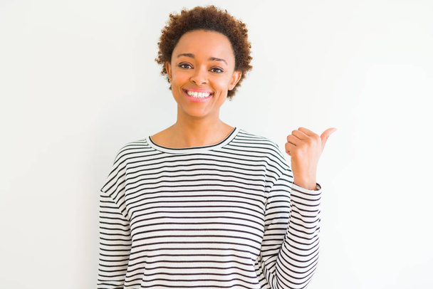 Молодая красивая африканская американка в полосатом свитере на белом фоне улыбается со счастливым лицом, глядя и указывая на сторону с большим пальцем вверх
. - Фото, изображение