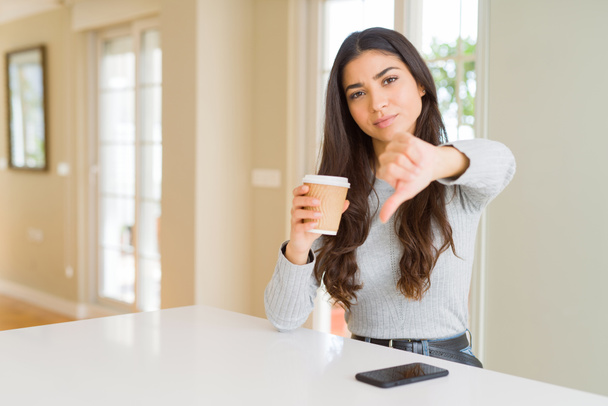 Молода жінка п'є чашку кави вдома з розлюченим обличчям, негативний знак, що показує нелюбов з великими пальцями вниз, концепція відторгнення
 - Фото, зображення