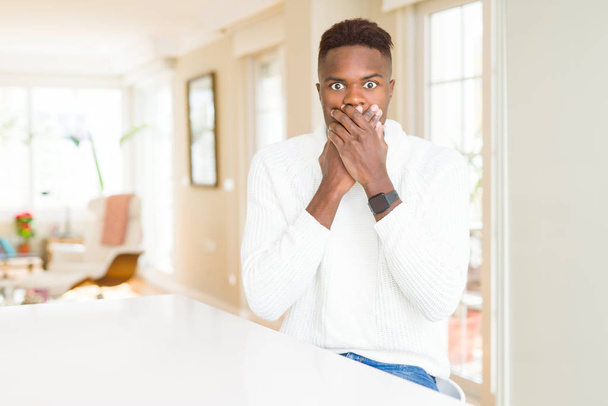 gutaussehender afrikanisch-amerikanischer Mann auf weißem Tisch schockiert, den Mund aus Versehen mit den Händen zu bedecken. Geheimes Konzept. - Foto, Bild