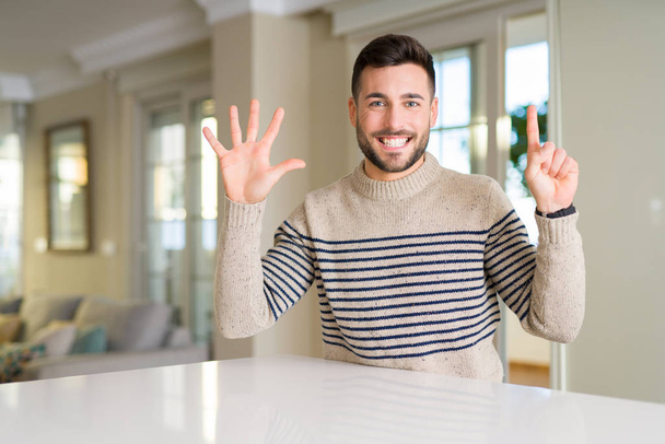 Όμορφος νεαρός στο σπίτι δείχνει και στραμμένο προς τα επάνω με τα δάχτυλα αριθμό έξι, ενώ χαμογελώντας αυτοπεποίθηση και χαρούμενοι. - Φωτογραφία, εικόνα