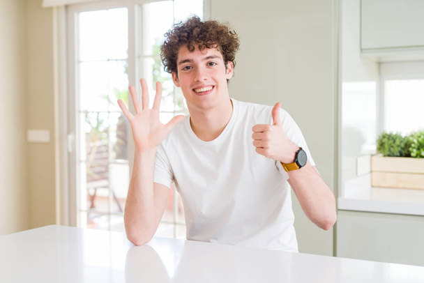 Joven hombre guapo con camiseta blanca en casa mostrando y apuntando hacia arriba con los dedos número seis mientras sonríe confiado y feliz
. - Foto, imagen