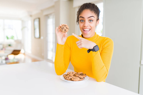 Young african american girl eating chocolate chips cookies as sweet snack very happy pointing with hand and finger - 写真・画像