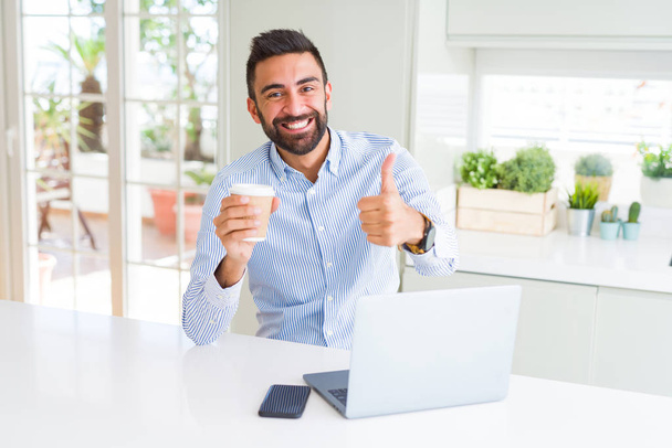 schöner hispanischer Mann arbeitet mit Computer-Laptop und trinkt eine Tasse Kaffee glücklich mit großem Lächeln tun ok Zeichen, Daumen hoch mit den Fingern, ausgezeichnetes Zeichen - Foto, Bild