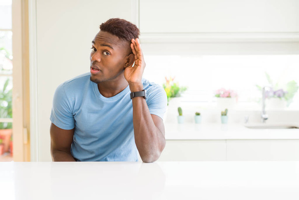 Hombre afroamericano guapo vistiendo camiseta casual en casa sonriendo con la mano sobre la oreja escuchando un rumor o chismes. Concepto de sordera
. - Foto, Imagen