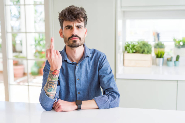Giovane uomo che indossa camicia casual seduto sul tavolo bianco Mostrando dito medio, maleducato e maleducato cazzo off espressione
 - Foto, immagini