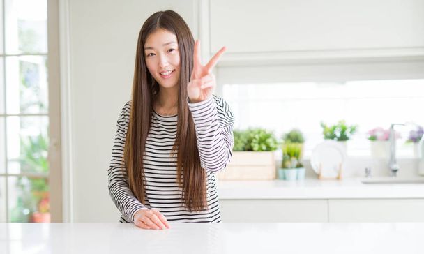 Gyönyörű ázsiai nő viselt csíkos pulóver mosolyogva, keres, hogy a kamera mutatja a győzelem jele ennek ujjak. Két szám. - Fotó, kép