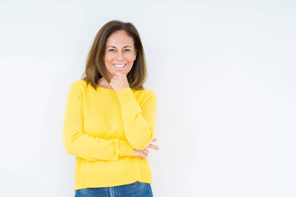 Bella donna di mezza età che indossa maglione giallo su sfondo isolato guardando fiducioso la fotocamera con il sorriso con le braccia incrociate e la mano sollevata sul mento. Pensare positivo
. - Foto, immagini
