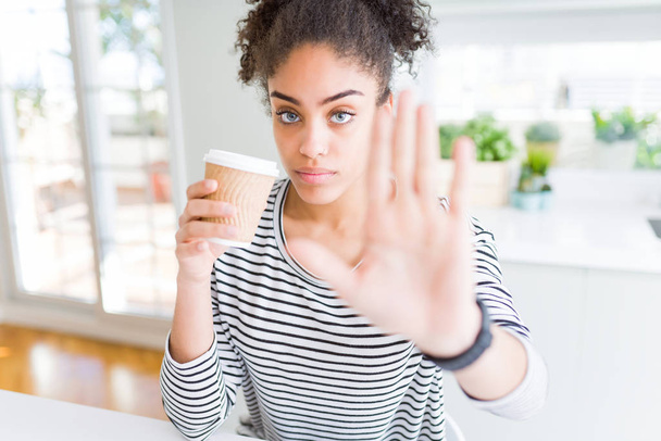 Jeune fille afro-américaine buvant un café sur une tasse en papier à emporter avec la main ouverte faisant stop signe avec expression sérieuse et confiante, geste de défense
 - Photo, image
