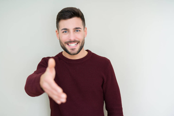 Jonge knappe man dragen een trui over geïsoleerde achtergrond lachende vriendelijke aanbod als begroeting en het welkom heten handdruk. Succesvol bedrijf. - Foto, afbeelding
