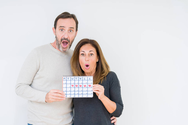Middle age couple holding month calendar over isolated background scared in shock with a surprise face, afraid and excited with fear expression - Foto, Imagem