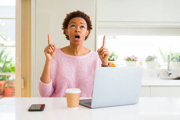 Mujer afroamericana joven que trabaja usando computadora portátil sorprendida y sorprendida mirando hacia arriba y señalando con los dedos y los brazos levantados
. - Foto, Imagen