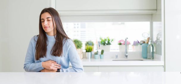 Širokoúhlý obraz krásné mladé ženy sedí na bílém stole doma s rukou na žaludek, protože nevolnost, bolestivé nemoci, pocit nevolnosti. Bolest koncept. - Fotografie, Obrázek