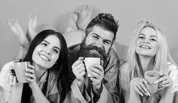 Lovers drinking coffee in bed. Man and women, friends on smiling faces lay, pink background. Man and women in domestic clothes, pajamas. Threesome relax in morning with coffee. Lovers in bed concept - Photo, Image
