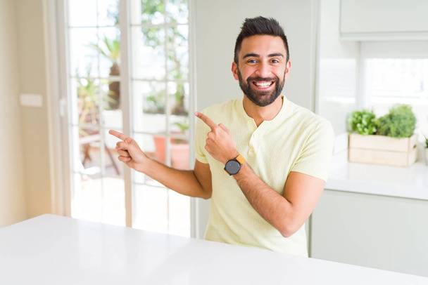Bonito homem hispânico casual camiseta amarela em casa sorrindo e olhando para a câmera apontando com duas mãos e dedos para o lado
. - Foto, Imagem